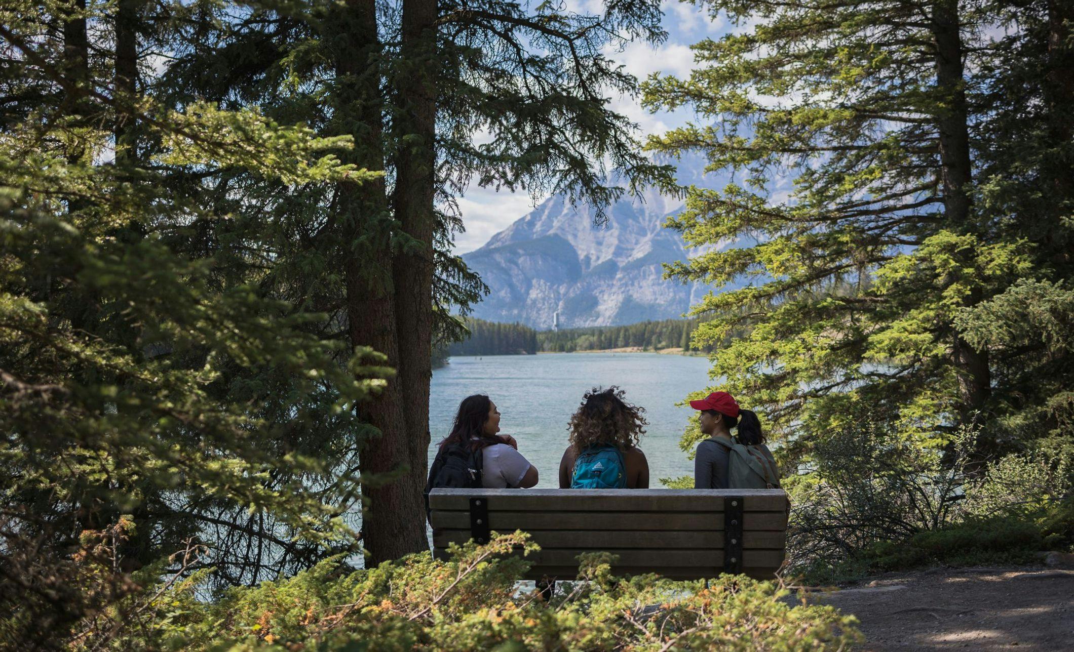 Three friends sit on a bench in a forested area looking out at a glacial blue lake